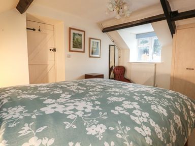 A bedroom with a bed with floral bedding a wooden door two framed pictures a red chair a standing mirror and a window at Poppy Cottage in Beaminster