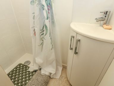 A bathroom corner with a sink cabinet a soap bar a shower with a floral curtain and a rubber mat on the floor at Poppy Cottage Beaminster