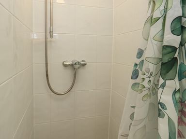 A tiled shower area with a silver showerhead and a shower curtain with green leaf patterns at Poppy Cottage in Beaminster