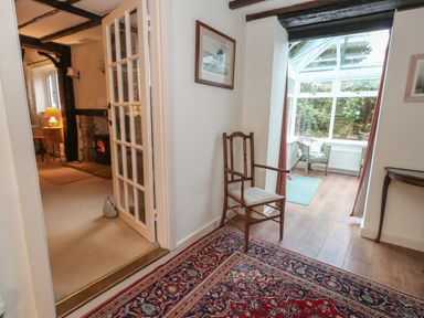 A hallway with a wooden chair and rug leading to a carpeted living room with a fireplace and a glass conservatory with chairs at Poppy Cottage in Beaminster