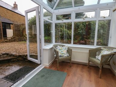 A conservatory with two wicker chairs with cushions a glass door opening to a garden at Poppy Cottage in Beaminster
