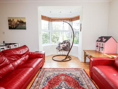 A living room with two red leather sofas a hanging chair by a bay window a wooden table with a pink dollhouse and a patterned rug on the floor at Trem Y Don Dolgarrog