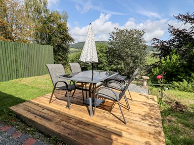 An outdoor wooden deck with a glass table and four chairs surrounded by grass and trees at Trem Y Don in Dolgarrog