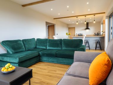 A living room with a green sectional sofa, a gray sofa with orange cushion, and a kitchen area with bar stools and hanging lights at Mynydd Y Glyn Lodge in Llanfyllin near Llanrhaeadr-Ym-Mochnant