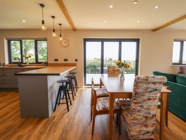 A dining area with a table and patterned chairs next to a kitchen island with stools and large windows at Mynydd Y Glyn Lodge in Llanfyllin near Llanrhaeadr-Ym-Mochnant