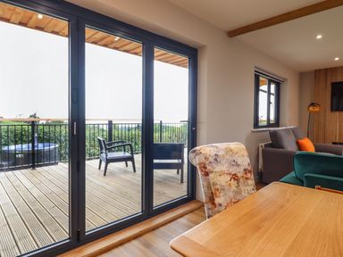 A living room with sliding glass doors leading to a wooden deck with chairs and a hot tub at Mynydd Y Glyn Lodge in Llanfyllin near Llanrhaeadr-Ym-Mochnant