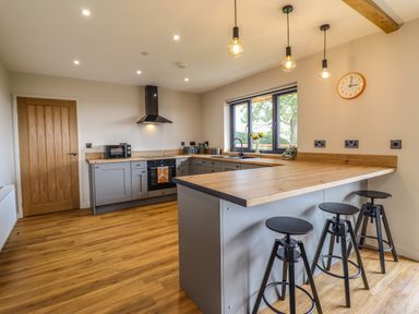 A kitchen with wooden floors grey cabinets a wooden countertop three black stools a window and a clock on the wall at Mynydd Y Glyn Lodge in Llanfyllin near Llanrhaeadr-Ym-Mochnant