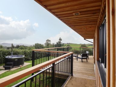 A wooden deck with black chairs and railing overlooking a garden with a hot tub at Mynydd Y Glyn Lodge in Llanfyllin near Llanrhaeadr-Ym-Mochnant