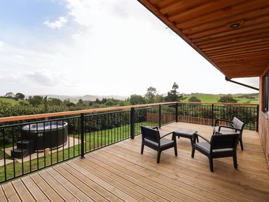 A wooden deck with black chairs and table overlooking a green landscape with a hot tub in the yard at Mynydd Y Glyn Lodge Llanfyllin near Llanrhaeadr-Ym-Mochnant