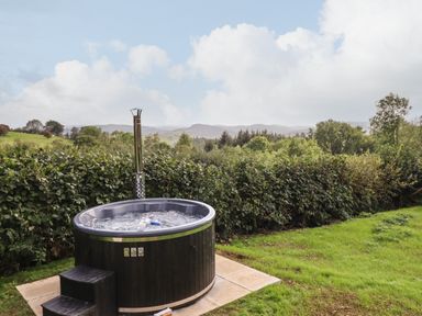 An outdoor hot tub on a paved area next to grass with trees and hills in the background at Mynydd Y Glyn Lodge in Llanfyllin near Llanrhaeadr-Ym-Mochnant