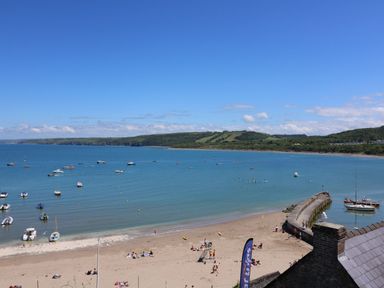 A beach with people, boats in the water, a pier, and green hills in the background at Towyn Hall in New Quay