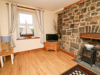 A room with a stone fireplace a small wood stove a TV on a wooden corner cabinet a window with striped curtains and a model sailboat on the windowsill at Towyn Hall in New Quay