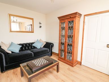 A living room with a black sofa with cushions a glass coffee table with a chess board a wooden cabinet with DVDs a large wall mirror and a white door at Towyn Hall in New Quay