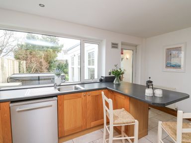 A kitchen with wooden cabinets a black countertop a stainless steel sink and dishwasher two white chairs and a window showing outside at Towyn Hall in New Quay