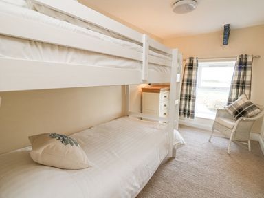 A bedroom with a white bunk bed a wooden chest of drawers a wicker chair with a cushion and a window with plaid curtains at Towyn Hall in New Quay