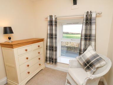 A room corner with a white chest of drawers with a table lamp on top and a white wicker chair with a plaid cushion near a window with plaid curtains at Towyn Hall in New Quay