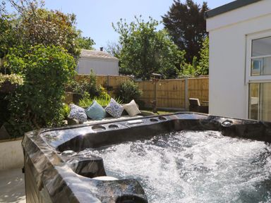 A bubbling hot tub next to a white building with a garden couch and pillows in a fenced backyard at Towyn Hall in New Quay