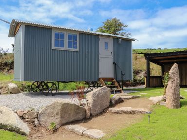 A small corrugated metal shepherds hut on wheels with a white door and window beside a gravel path and large rocks at Bosulla Shepherds Hut in Penzance