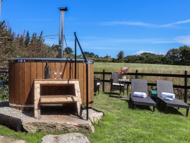 An outdoor area with a wooden hot tub with steps two lounge chairs with towels a small table with flowers and chairs in a grassy fenced field at Bosulla Shepherds Hut in Penzance