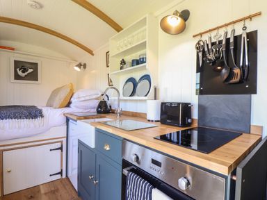 A compact kitchen area with sink stove oven and utensils next to a small bed with pillows and blanket at Bosulla Shepherds Hut in Penzance