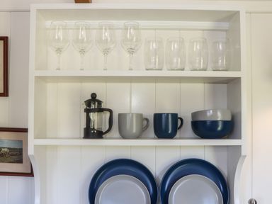 A kitchen shelf with wine glasses, drinking glasses, a French press, mugs, bowls, and plates at Bosulla Shepherds Hut in Penzance