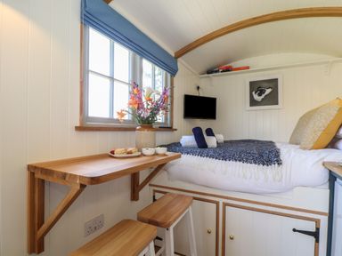 A small bedroom with a wooden fold-down table flowers on the table stools and a bed with cushions at Bosulla Shepherds Hut in Penzance