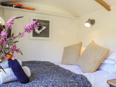 A bedroom with a made bed with cushions and folded towels and flowers on a side table at Bosulla Shepherds Hut in Penzance