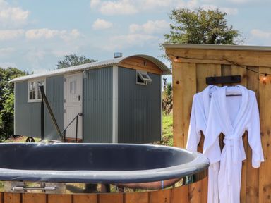 An outdoor hot tub with two white robes hanging on a wooden wall and a small gray shepherds hut in the background at Bosulla Shepherds Hut in Penzance
