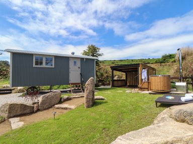 A small corrugated metal hut with steps leading to the door next to a wooden covered seating area and a wooden hot tub on grass at Bosulla Shepherds Hut in Penzance