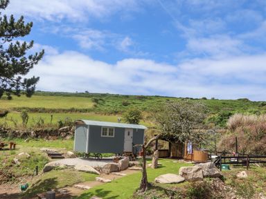 A small blue shepherds hut on wheels in a rural grassy area with rocks trees and a wooden seating area at Bosulla Shepherds Hut in Penzance