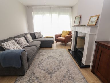 A living room with a grey sectional sofa with cushions a brown armchair a fireplace with framed art and a wooden cabinet at 19A Lighthouse Village in Fenit County Kerry