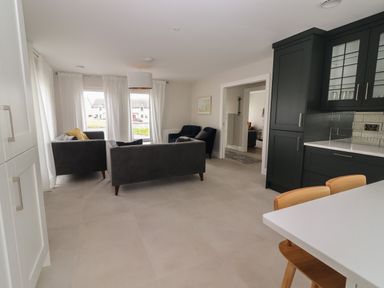A living area with grey sofas and armchairs near large windows and dark kitchen cabinets at 19A Lighthouse Village in Fenit County Kerry
