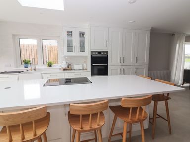 A kitchen with a white island and wooden stools near cabinets and windows at 19A Lighthouse Village in Fenit County Kerry