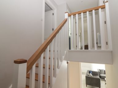 A staircase with wooden handrail and white balusters leading to a hallway with doors and a view of a living room with grey sofas at 19A Lighthouse Village in Fenit County Kerry