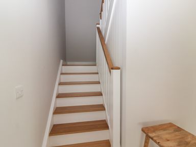 A wooden staircase with white risers and a wooden handrail next to a white wall with a light switch and a wooden stool at 19A Lighthouse Village in Fenit County Kerry