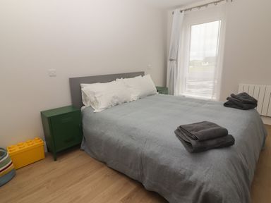 A bedroom with a bed covered in gray bedding and white pillows two green bedside cabinets a window with white curtains and folded towels on the bed at 19A Lighthouse Village in Fenit County Kerry