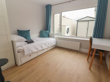 A bedroom with a white bed with teal pillows a desk and chair next to a large window at 19A Lighthouse Village in Fenit County Kerry