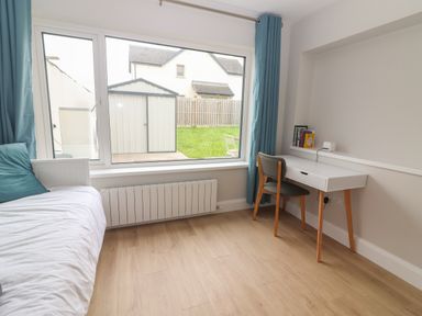 A bedroom with a bed, desk, chair, and a large window showing a shed and garden at 19A Lighthouse Village in Fenit County Kerry
