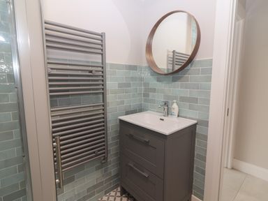 A bathroom with a gray vanity cabinet with sink a wall-mounted round mirror and a gray towel radiator at 19A Lighthouse Village in Fenit County Kerry