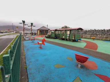An outdoor children's playground with a toy train, spring riders, a swing set, and a stone wall in the background at 19A Lighthouse Village in Fenit County Kerry