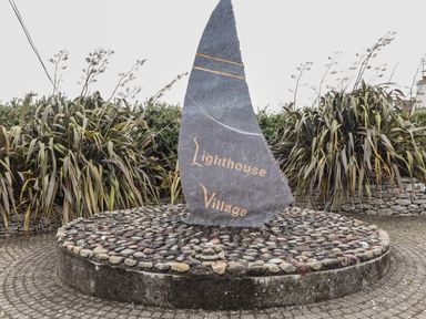 A stone monument shaped like a sail with lighthouse village text on a circular stone base surrounded by plants at 19A Lighthouse Village in Fenit County Kerry