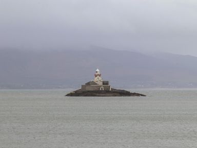 A lighthouse on a small rocky island in the sea with mountains and a cloudy sky in the background at 19A Lighthouse Village in Fenit County Kerry