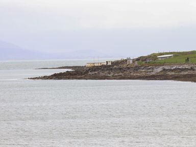 A coastal scene with rocky shore grassy hill and small structures near the water at 19A Lighthouse Village in Fenit County Kerry