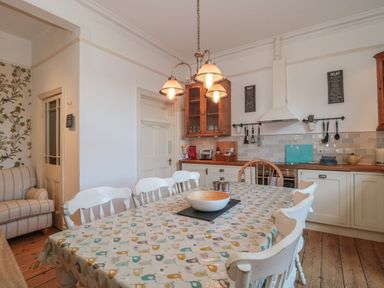 A kitchen with a wooden table covered with a patterned tablecloth surrounded by white chairs and wooden cabinets at 49 Esplanade in Burnham-On-Sea