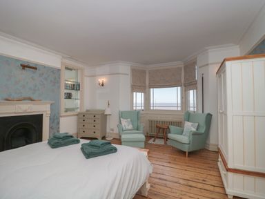 A bedroom with a bed, two armchairs, a chest of drawers, a wardrobe, and windows overlooking the sea at 49 Esplanade in Burnham-On-Sea