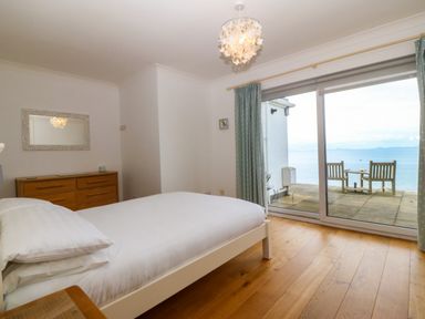 A bedroom with a bed a wooden dresser a hanging light and a sliding door to a patio with chairs overlooking the sea at Gwylanod Llwyngwril near Fairbourne