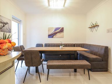 A dining area with a wooden table surrounded by brown upholstered bench seating and chairs with wall art and a fruit bowl at Gwylanod Llwyngwril near Fairbourne
