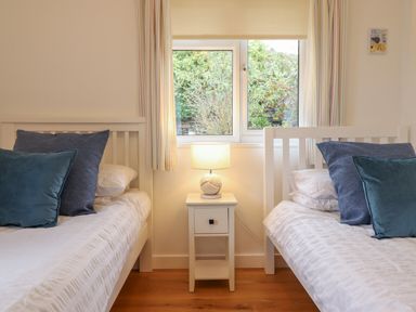 A bedroom with two white beds separated by a white nightstand with a lamp in front of a window at Gwylanod in Llwyngwril near Fairbourne