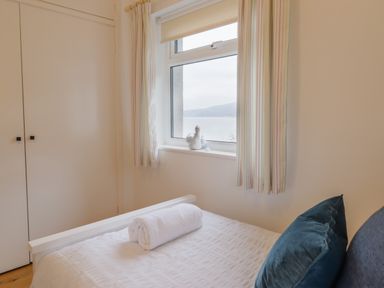 A bedroom with a bed with white bedding and a rolled towel a window with curtains and a small decoration on the sill at Gwylanod Llwyngwril near Fairbourne