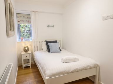 A small bedroom with a bed with pillows and towel a bedside table with a lamp and a window with a decorative blind and curtain at Gwylanod in Llwyngwril near Fairbourne
